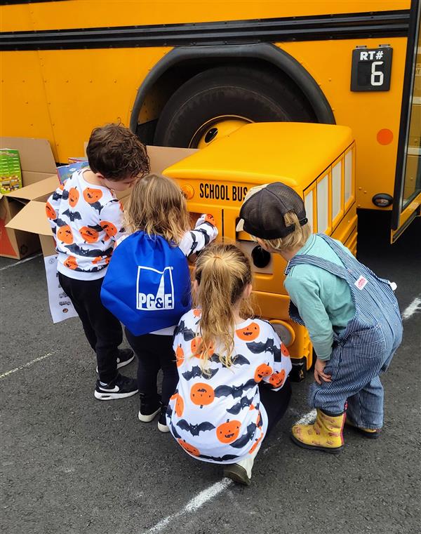 Four kids crouched in front of a large toy school bus sitting outside of a real school bus.