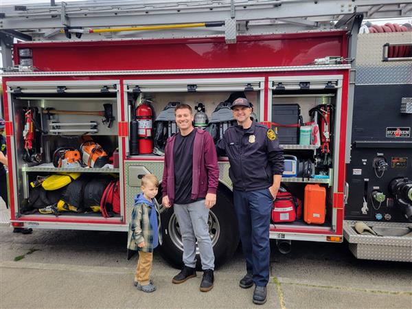 Firefighter, man, and boy smile in front of a firetruck. 