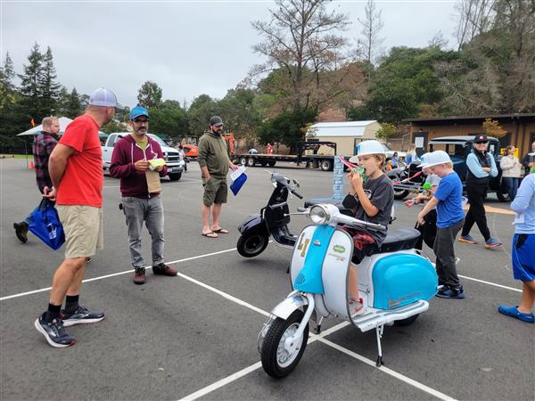 Boy sitting on a vespa eating an ice cream wearing a plastic hard hat at a school event.